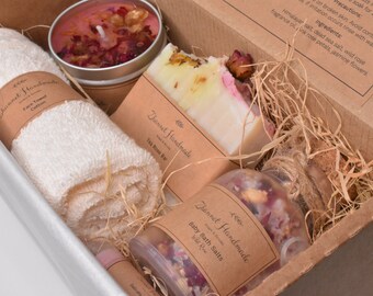 Puede incluir: Una caja de regalo llena de productos para el baño y el cuerpo. La caja contiene una toalla blanca, una pastilla de jabón rosa y blanca, un tarro de sales de baño, una vela rosa y un pequeño tubo de bálsamo labial. La caja está forrada con papel marrón y paja.