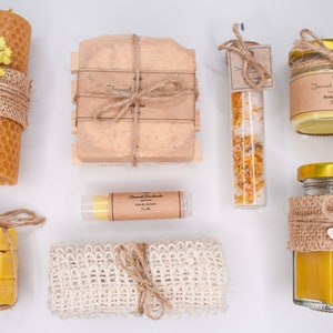 May include: A spa gift set featuring a honeycomb candle, a bar of soap, a lip balm, a bath scrub, a jar of body butter, and a jar of bath salts. The set is wrapped in hessian and twine and decorated with dried flowers.