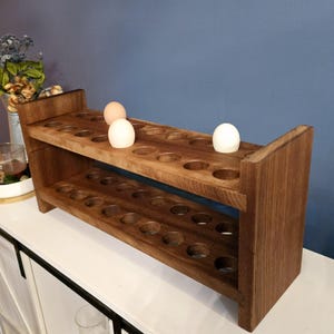 May include: A two-tiered, wooden egg holder with a rich brown finish. The holder has circular cutouts to hold eggs, with three eggs visible. The design is rustic and functional, perfect for kitchen storage. The holder is made of wood.