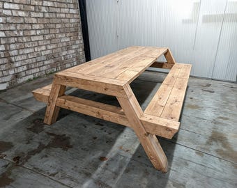 May include: A rustic wooden picnic table with two benches attached. The table is made of natural wood with a light brown finish. The table is in a warehouse setting with a brick wall in the background.