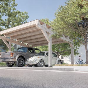Op de afbeelding: Een houten carport biedt beschutting aan twee oldtimers: een grijze pick-up truck en een witte Volkswagen Kever. De carport heeft een lichtbruin houten frame. Bomen en een wit gebouw completeren de scène.