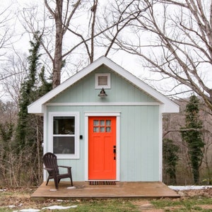 May include: A small, light blue house with a bright orange door and a wooden porch. The house is surrounded by trees.
