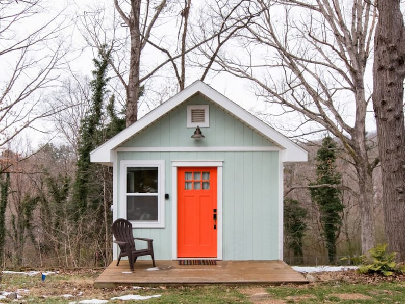 May include: A small, light blue house with a bright orange door and a wooden porch. The house is surrounded by trees.