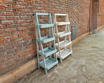 Puede incluir: Dos estanterías de madera en forma de escalera, una pintada de azul claro y la otra de blanco, se apoyan en una pared de ladrillo. Ambas estanterías tienen cuatro niveles.