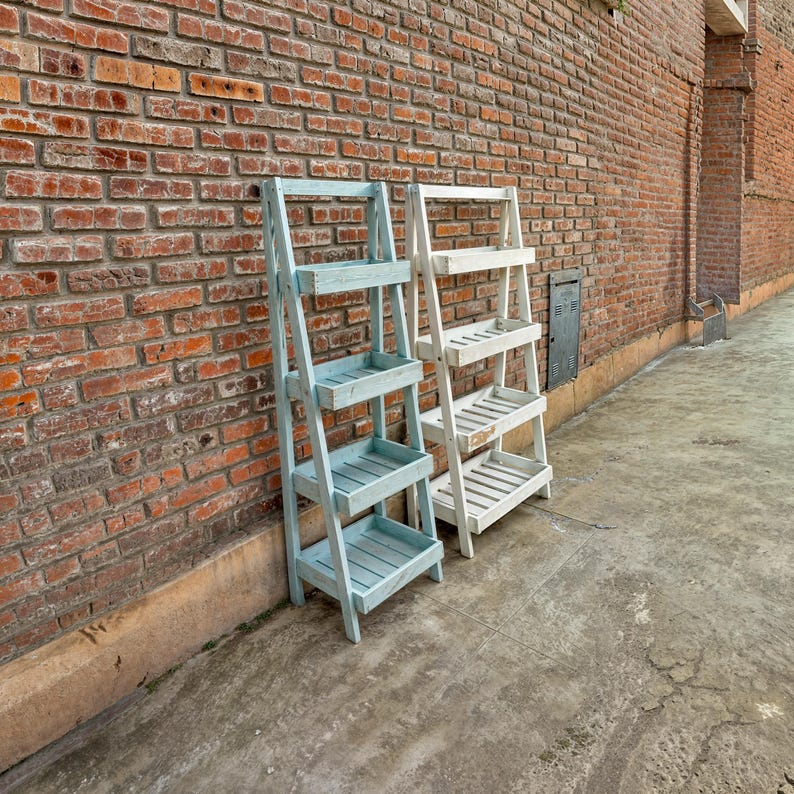 May include: Two wooden ladder shelves, one painted light blue and the other white, stand against a brick wall. Both shelves have four tiers.