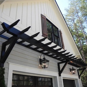 Puede incluir: Una pérgola negra con un marco de madera y un diseño de celosía está unida al lado de una casa blanca y gris. La pérgola está sostenida por dos soportes negros y tiene dos faroles negros colgando de ella.