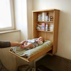 Peut inclure: Une table à langer en bois avec un matelas à langer rabattable et une étagère au-dessus pour ranger les accessoires de bébé. Un bébé est allongé sur le matelas à langer, et une main se tend pour toucher le pied du bébé.