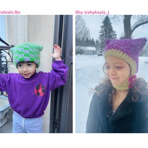 May include: Two children wearing handmade cat ear hats. One hat is green and purple, the other is purple and green. The child on the left is wearing a purple sweatshirt with a graphic. The child on the right is wearing a black coat.