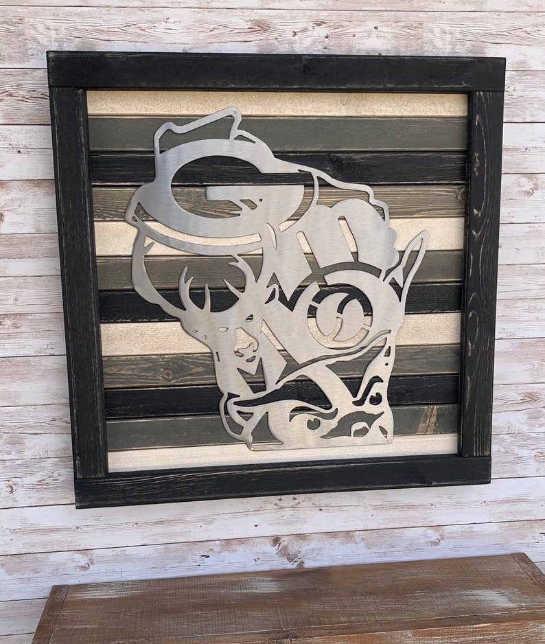 K&ouml;nnte beinhalten: Ein Metallausschnitt des Bundesstaates Wisconsin mit einem Hirsch und dem Wort "G-MO" darin. Der Ausschnitt ist auf einem Holzrahmen mit einem mehrfarbigen Holzmaserungsmuster montiert.