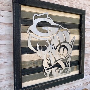 K&ouml;nnte beinhalten: Ein gerahmtes Wandbild aus Metall mit einer Hirsch-Silhouette, in deren Geweih ein stilisiertes "G" und "M" eingearbeitet sind. Der Hintergrund ist ein mehrfarbiger Holzplanken-Design in Schwarz-, Wei&szlig;- und Graut&ouml;nen.