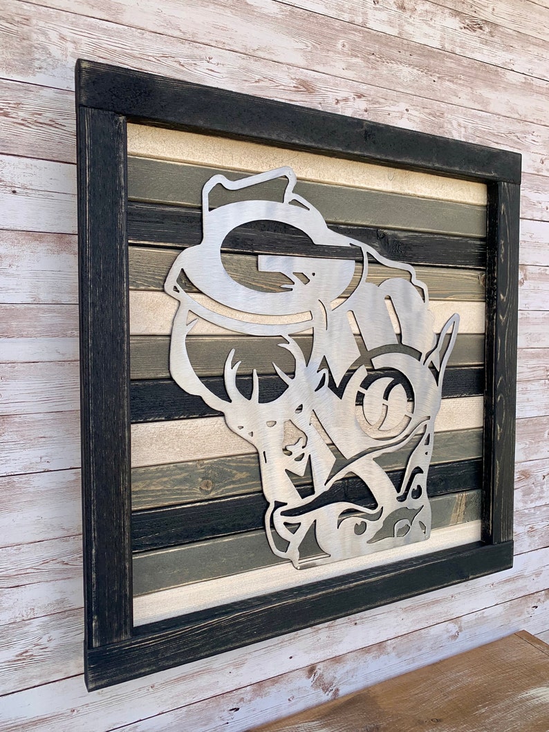 K&ouml;nnte beinhalten: Ein gerahmtes Wandbild aus Metall mit einer Hirsch-Silhouette, in deren Geweih ein stilisiertes "G" und "M" eingearbeitet sind. Der Hintergrund ist ein mehrfarbiger Holzplanken-Design in Schwarz-, Wei&szlig;- und Graut&ouml;nen.