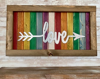 May include: A rectangular wooden sign with a rainbow-colored background and a brown frame. The word "love" is written in silver script with an arrow through it. The background is made of vertical wooden planks painted in various colors.