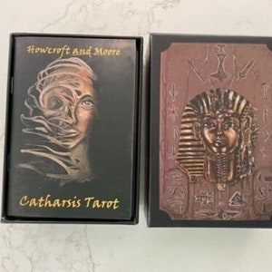 May include: Two black boxes with gold lettering. The box on the left has a black and white image of a woman's face with a swirling design. The text on the box reads "Howcroft and Moore Catharsis Tarot". The box on the right has a gold image of a pharaoh's head with a gold background.