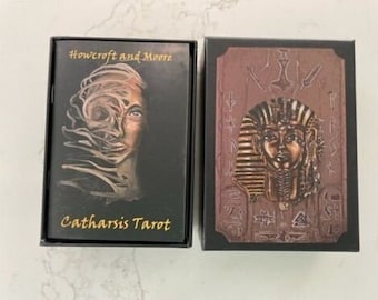 May include: Two black boxes with gold lettering. The box on the left has a black and white image of a woman's face with a swirling design. The text on the box reads "Howcroft and Moore Catharsis Tarot". The box on the right has a gold image of a pharaoh's head with a gold background.