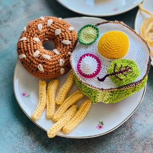 Puede incluir: Un juego de desayuno de ganchillo en un plato blanco con detalles florales. El juego incluye un bagel, huevo frito, papas fritas y otros alimentos. El bagel es marrón con detalles blancos. El huevo frito tiene una yema amarilla y detalles verdes y rosas.