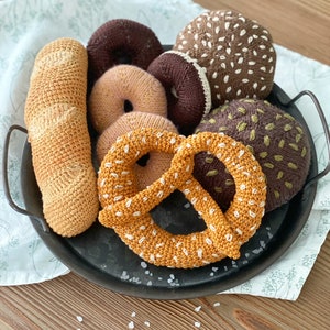 Peut inclure: Une collection d'aliments en crochet, dont un bretzel, une baguette, un beignet et deux types de petits pains, tous disposés sur un plateau en métal noir.