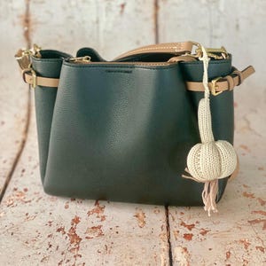 May include: Dark green handbag with tan accents, including straps and buckles. The bag has a textured appearance and a zippered top. A decorative, knitted charm hangs from the side. The bag is on a distressed wooden surface.