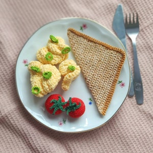 Puede incluir: Un juego de desayuno tejido a crochet en un plato blanco con un patrón floral. El juego incluye una pieza de tostada en forma de triángulo, tres piezas de huevos revueltos y dos tomates rojos.