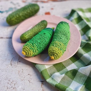 May include: Three crocheted green and yellow pickles on a pink plate with a green and white checkered tablecloth.