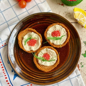 Pode incluir: Um prato de cerâmica marrom contém três porta-copos de sushi de crochê. Cada porta-copos apresenta uma borda marrom, um centro branco com detalhes de contas vermelhas e detalhes de fio verde. Tomates cereja e outros alimentos de crochê estão no fundo.