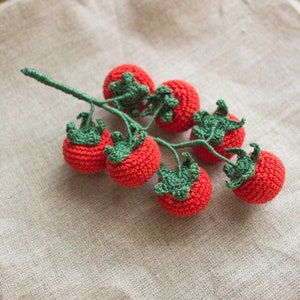 Op de afbeelding: Een gehaakte tomatenplant met zes rode tomaten en groene bladeren. De plant ligt op een beige stoffen achtergrond.