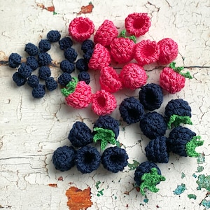 May include: A collection of handmade crochet berries in shades of pink and navy blue. The berries include raspberries and blueberries, with green leaves. The items are arranged on a distressed white surface.
