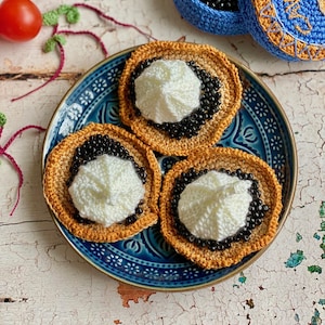 Pode incluir: Um prato de cerâmica azul contém três itens de comida de crochê com um centro branco, bordas de contas pretas e bordas bege. Dois tomates vermelhos e um recipiente azul de crochê com contas pretas também são visíveis.