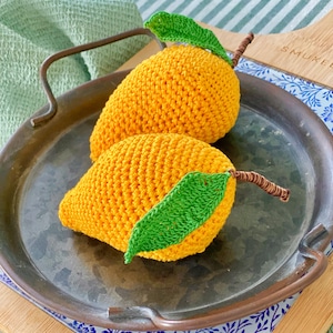 Op de afbeelding: Twee handgemaakte gehaakte mango's in helder geel met groene bladeren en bruine stelen. De mango's liggen op een metalen dienblad met handvatten, op een patroonachtergrond. Het haakwerk is gedetailleerd en getextureerd.