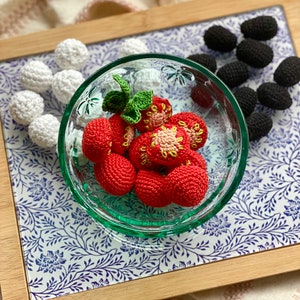 Könnte beinhalten: Eine Glasschüssel gefüllt mit gehäkelten roten Erdbeeren mit grünen Blättern. Die Schüssel steht auf einem Holztablett mit einem blauen und weißen Blumenmuster. Um die Schüssel herum liegen gehäkelte weiße und schwarze Kugeln.