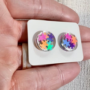 May include: A pair of clear resin stud earrings with colorful puzzle pieces inside. The earrings are on a white card.