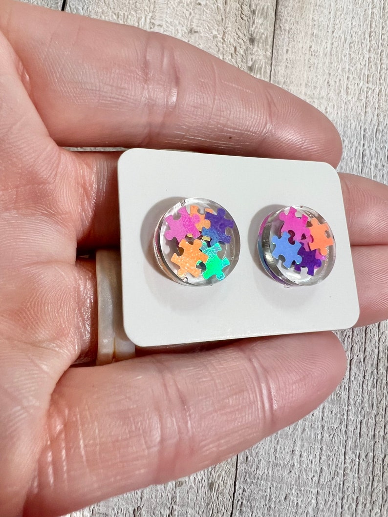 May include: A pair of clear resin stud earrings with colorful puzzle pieces inside. The earrings are on a white card.