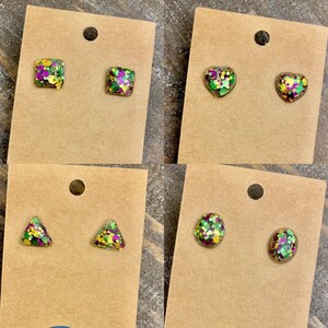 May include: Four pairs of stud earrings with different shapes, including square, heart, triangle, and circle. Each earring is filled with green, purple, and gold glitter.