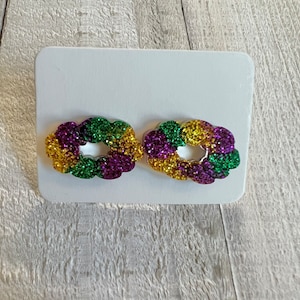 May include: A pair of purple, green, and gold glitter earrings shaped like a Mardi Gras king cake.