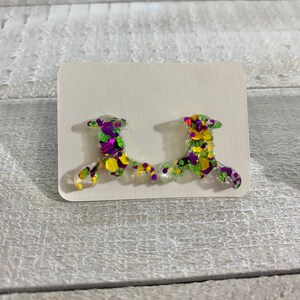 May include: A pair of clear, plastic earrings shaped like a Mardi Gras mask. The earrings are decorated with green, purple, and gold glitter.