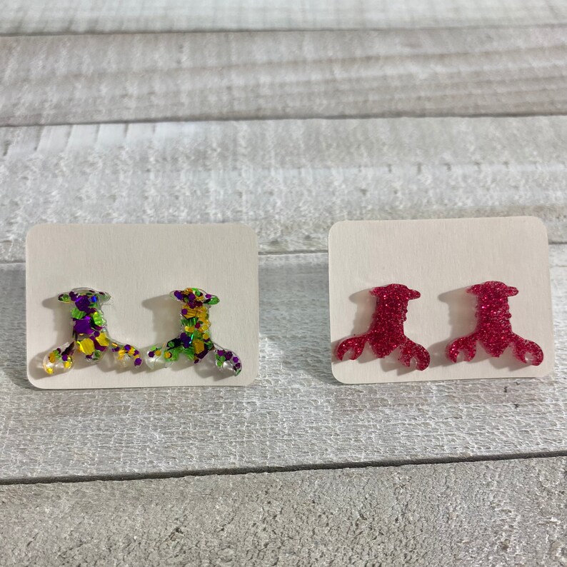 May include: Two pairs of stud earrings. One pair is red glitter and the other pair is green, purple, and gold glitter.
