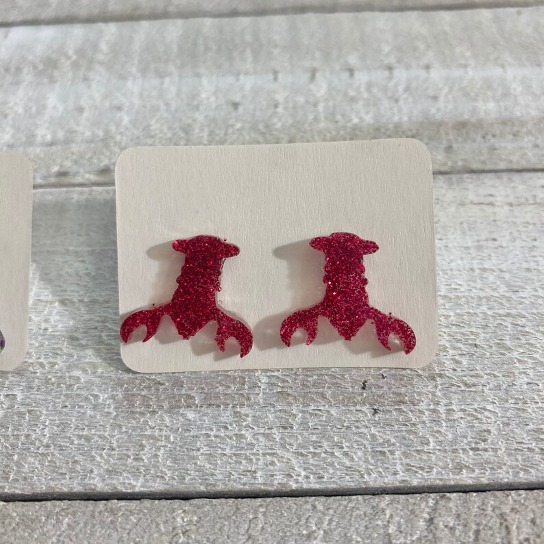 May include: A pair of pink glitter lobster earrings. The earrings are shaped like a lobster claw and are made of a sparkly pink material.