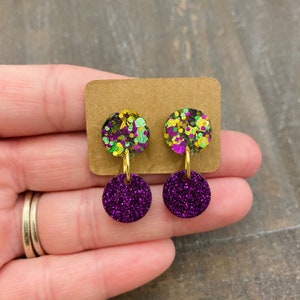 May include: A pair of dangle earrings with two-toned round discs. The top disc is green, gold, and purple glitter. The bottom disc is solid purple glitter.