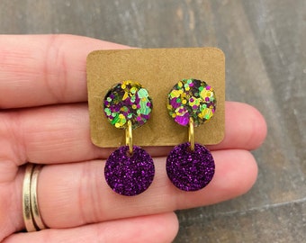Op de afbeelding: Een paar oorbelletjes met twee ronde schijven. De bovenste schijf is bedekt met groene, gouden en paarse glitter. De onderste schijf is bedekt met paarse glitter.