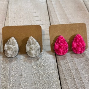 May include: Two pairs of teardrop-shaped stud earrings. One pair features white, crystal-like stones with a silver-coloured border. The other pair showcases bright pink, crystal-like stones with a silver-coloured border. Both sets are displayed on brown earring cards.