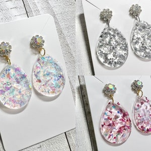 May include: Three pairs of dangle earrings with clear teardrop-shaped resin that has iridescent glitter embedded in it. The tops of the earrings are adorned with rhinestones.