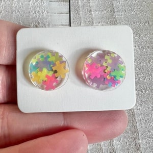 May include: A pair of clear resin stud earrings with colorful puzzle pieces inside. The earrings are on a white card.