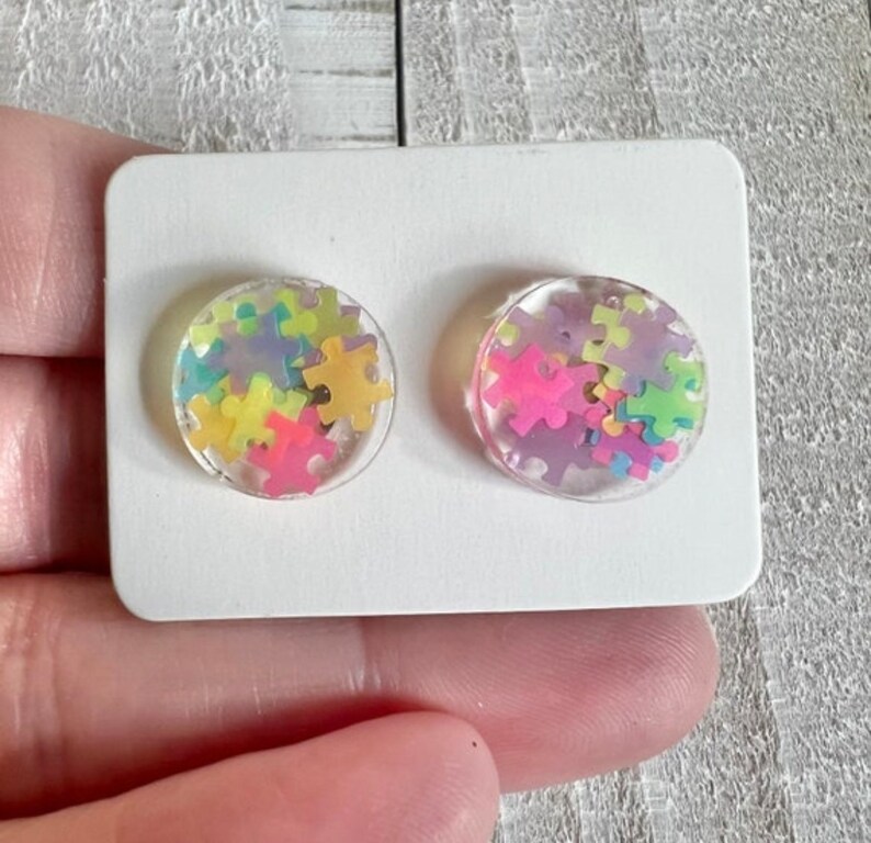 May include: A pair of clear resin stud earrings with colorful puzzle pieces inside. The earrings are on a white card.