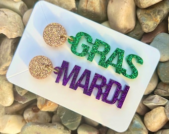 May include: A pair of Mardi Gras earrings with gold glitter accents. One earring features the word "GRAS" in green glitter and the other earring features the word "MARDI" in purple glitter.