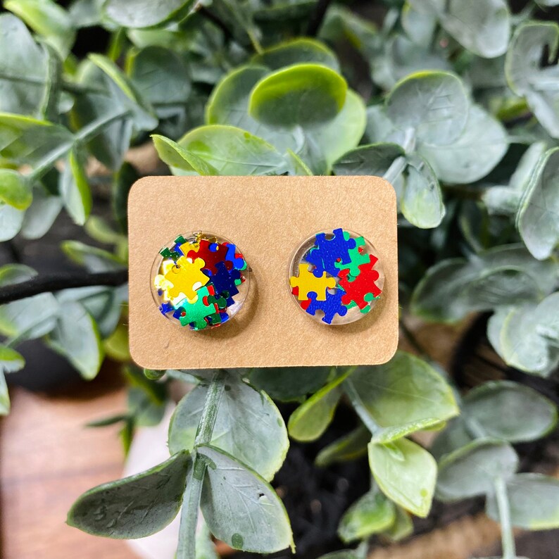 May include: A pair of round stud earrings with a clear resin base filled with colorful puzzle pieces. The puzzle pieces are in shades of blue, green, yellow, and red.