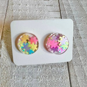 May include: A pair of round, clear resin earrings with colorful puzzle piece confetti inside. The earrings are on a white card.