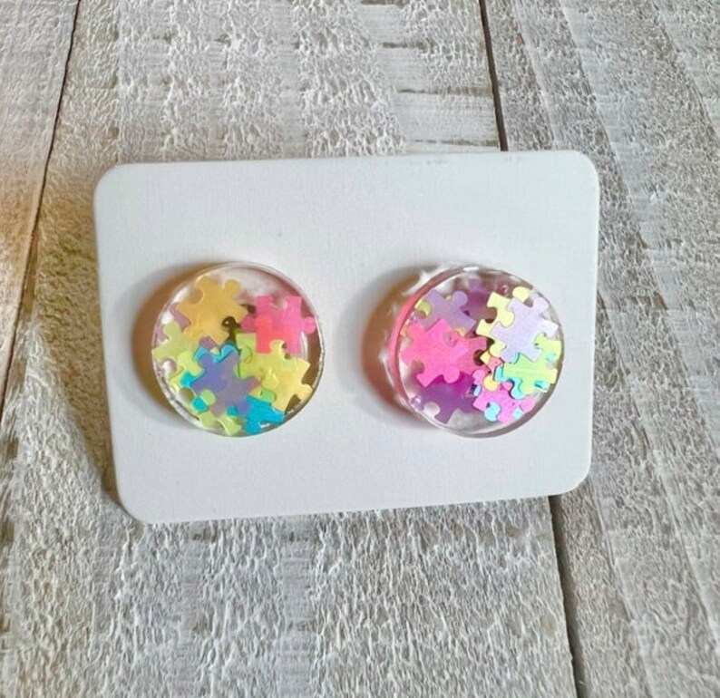 May include: A pair of round, clear resin earrings with colorful puzzle piece confetti inside. The earrings are on a white card.