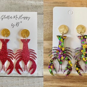May include: Two pairs of dangle earrings featuring a lobster design. One pair is red glitter with gold accents, the other is green, purple, and gold glitter. The earrings are on white cards with the text "Glitter Me Happy by KV".