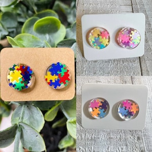 May include: Three pairs of stud earrings with a clear resin base and colorful puzzle pieces inside. The puzzle pieces are in shades of blue, green, yellow, red, pink, and purple.