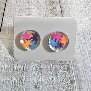 May include: A pair of stud earrings with a clear resin base and colorful puzzle pieces inside. The earrings are displayed on a white card.