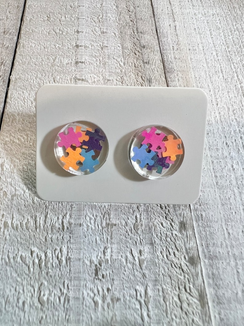 May include: A pair of stud earrings with a clear resin base and colorful puzzle pieces inside. The earrings are displayed on a white card.